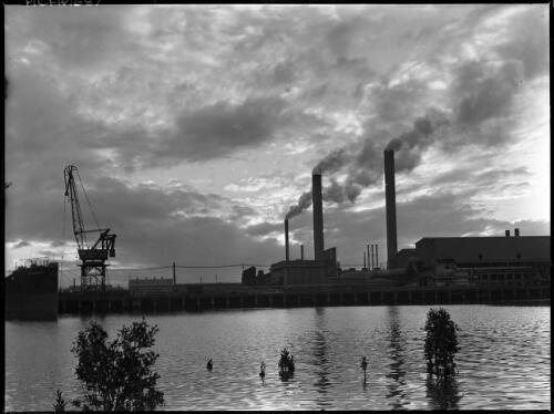 [River and a wharf in front of a factory with two smoke stacks and a crane, and the bow of the Kekerangu] [picture] : [Industry] / [Frank Hurley]
