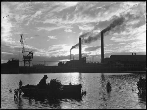 [Man in a rowing boat on a river in front of a wharf and a factory with two smoke stacks and a crane, and the bow of the Kekerangu, 1] [picture] : [Industry] / [Frank Hurley]