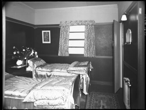[Bedroom with two single beds, heater, mirror, shelf, vase of flowers, mat, window and curtains, table and a door] [picture] / [Frank Hurley]