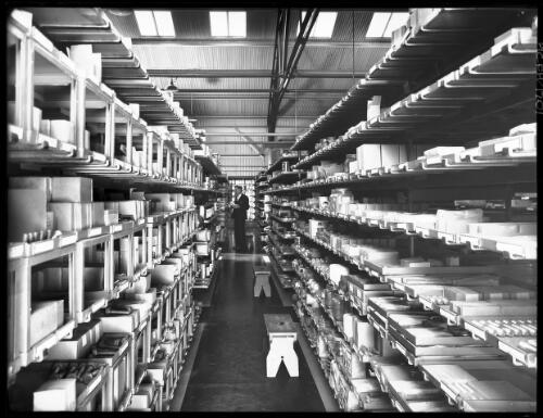 [Industrial shelves stacked with components in a factory or plant with stools and one figure wearing a suit] [picture] : [Industry] / [Frank Hurley]