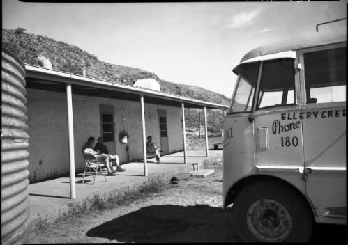 Accommodation Glen Helen [three figures sitting on a verandah near a tank and a bus with "Ellery Creek, Phone 180" painted on the side] [picture] : [Central Australia] / [Frank Hurley]