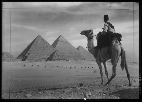 [The Great Pyramids with a figure on camel in foreground and column of camels with riders] [picture] : [Cairo, Egypt panoramas] / [Frank Hurley]