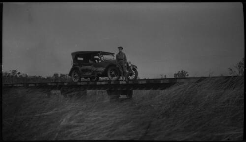 Sturts Plains, Newcastle [motor vehicle with two figures, on a low bridge, with long grass] [picture] : [Central Australia] / [Frank Hurley]