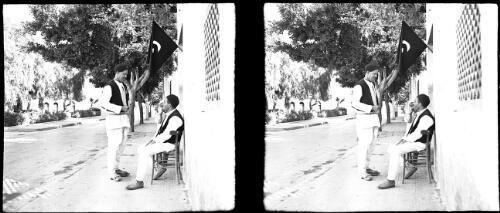 Derna [two men talking, one seated, a flag above them] [picture] : [Libya, World War II] / [Frank Hurley]