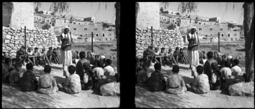 School at Bethany [children seated around teacher, a stone wall and buildings on a hillside in the background] [picture] / [Frank Hurley]