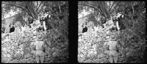 [Figure in pith helmet taking grapes, stone wall, figures up above] [picture] / [Frank Hurley]