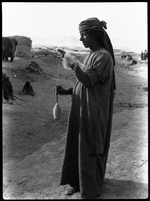 [A figure spinning wool on a small hand tool in the open air, other figures in the background, 1944] [picture] : [Iraq, World War II] / [Frank Hurley]