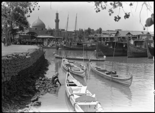 [Canoes in a canal with buildings on one side, including a minaret and a dome, with a rock wall, 1944] [picture] : [Iraq, World War II] / [Frank Hurley]