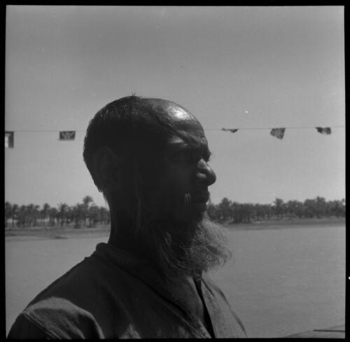 Chubaish [a bald man with a beard, facing right, in front of a string of flags and a river, 1944] [picture] : [Iraq, World War II] / [Frank Hurley]