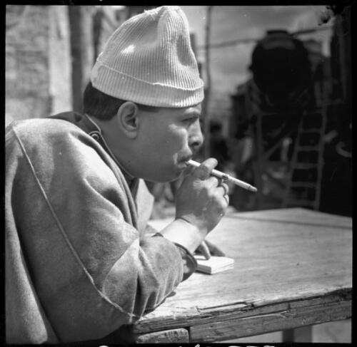 Village scenes shot at Misr studies on set [a portrait of a man wearing a knitted hat, leaning on a table, holding a cigar and a small box] [picture] : [Cairo, Egypt, World War II] / [Frank Hurley]