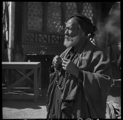 Village scenes shot at Misr, studies on set [portrait of a man wearing Arab robes, carrying a string of beads and with a table and four arched windows in the background] [picture] : [Cairo, Egypt, World War II] / [Frank Hurley]