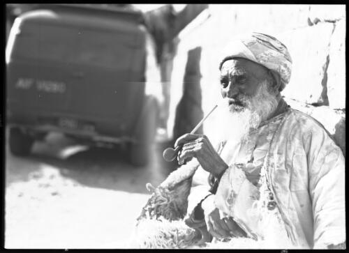 Jerusalem [figure of a man sitting with a pipe, leaning on a stone wall, and vehicle marked 'AIF V1280'? in background, Palestine] [picture] : [Portrait Studies, Libya, World War II] / [Frank Hurley]