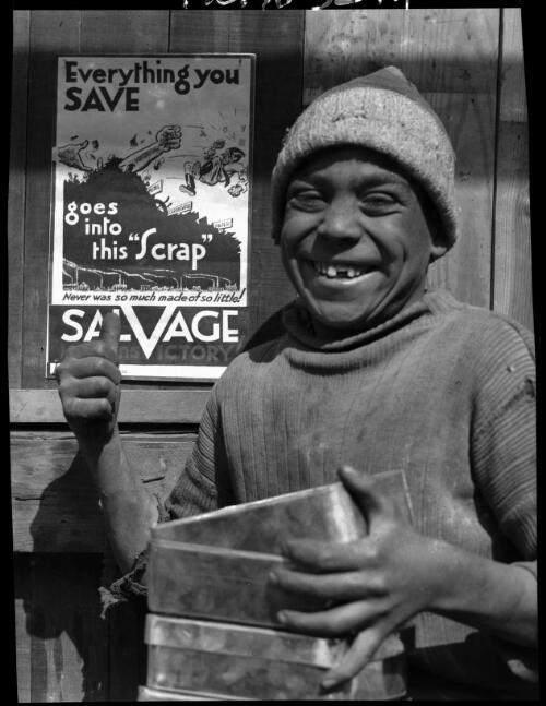 [Boy holding tins and pointing with his thumb at a poster, 'Everything you save goes into this Scrap. Never was so much made of so little! Salvage Hastens Victory!'] [picture] : [Portrait Studies, Libya, World War II] / [Frank Hurley]