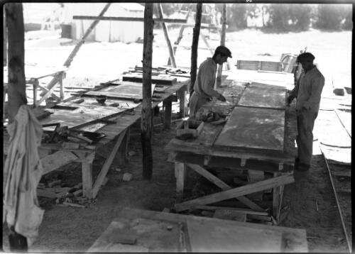 Jewish industry Qibet (Hayren?) [men working at tables in an outdoor covered area, scraping paint, with railway tracks at side of work area] [picture] / [Frank Hurley]