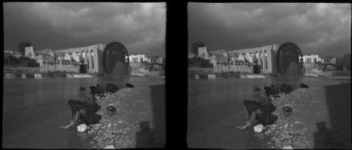 Hama, central Syria [view from riverbank across to waterwheel, aqueduct and picturesque townscape with seven figures at water's edge] [picture] : [Syria, World War II] / [Frank Hurley]