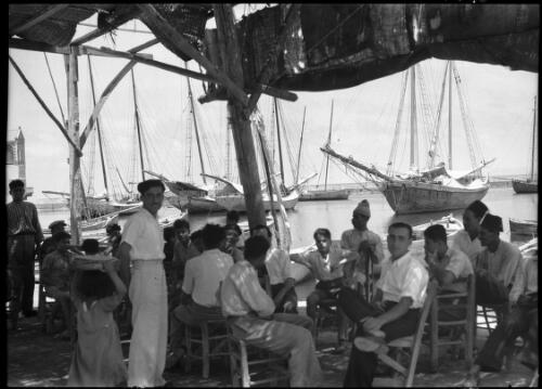 By the Port of Ancient Tyre [group of men seated on chairs, with fishing vessels behind] [picture] / [Frank Hurley]