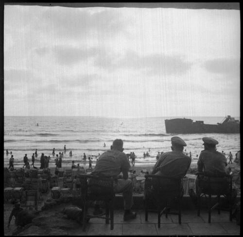 [Three army men on verandah look out to beach scene and ship] [picture] / [Frank Hurley]