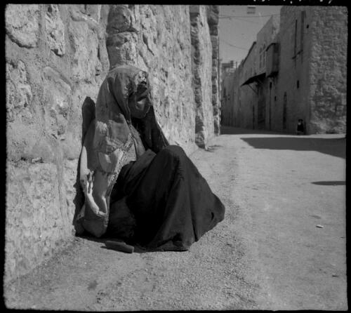 [Downcast woman with beaded veil sits leaning against street wall, Old City, Jerusalem (?)] [picture] / [Frank Hurley]
