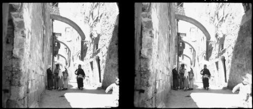 Sundry stereos, Jerusalem [view down narrow cobbled street, including Franciscan monk with umbrella, armed figure, two women and a child, in the Old City] [picture] / [Frank Hurley]