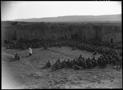 Tocra [large group of Australian soldiers sit listening to a speaker, building and camera crew behind, ca. 1940-1946] [picture] : [Barqah, Libya] / [Frank Hurley]