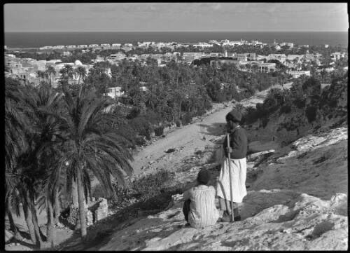 [Town on the ocean with two boys in foreground, dirt road and palm trees in between] [picture] : [Barqah, Libya] / [Frank Hurley]