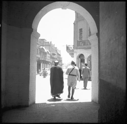 [View through an archway onto a building lined street, with two figures, one in robe and headdress and the other in uniform, World War II] [picture] : [Iran] / [Frank Hurley]