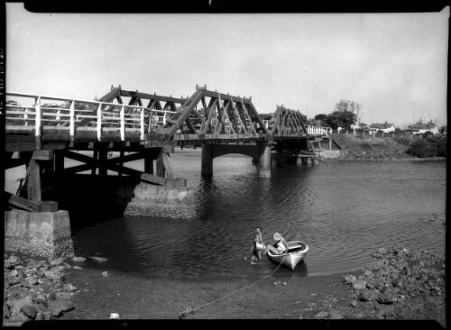 [Three people by a wooden boat, a bridge over the river forms a backdrop] [picture] / [Frank Hurley]