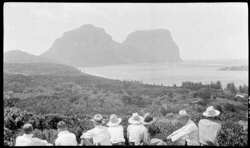 [Eight unidentified people sitting and looking at Mount Lidgbird and Mount Gower, Lord Howe Island] [picture] / [Frank Hurley]