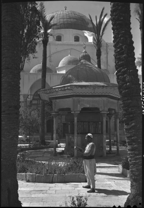 [Mosque with man standing and reading a book by small shrine, Acre, Palestine] [picture] / [Frank Hurley]