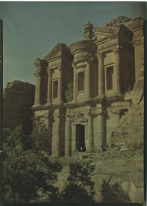 [Monastery (Al-Deir), Petra, with a standing figure at entrance and trees in the foreground] [picture] : [Jordan, World War II] / [Frank Hurley]