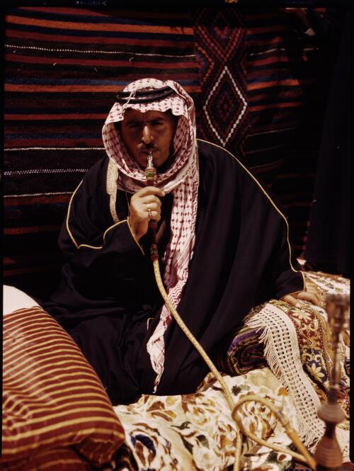 [Man in Arab dress sitting, smoking a pipe, with a carpet backdrop] [picture] : [Sudan, 1940's] / [Frank Hurley]