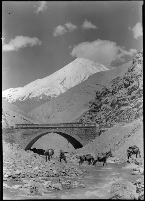 A man and four horses crossing a river next to a bridge with Mount Demavend in the background, Iran, ca. 1943 [picture] / [Frank Hurley]