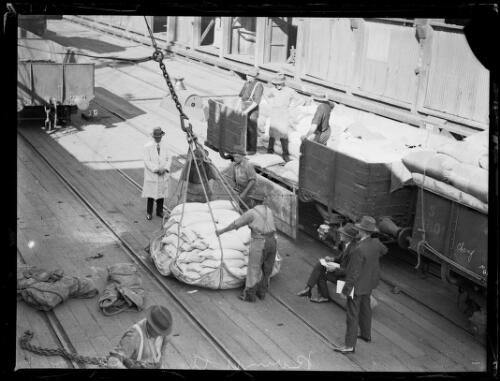 Workers using a sling crane to load goods from wagons on the wharf onto a ship, New South Wales, ca. 1920s [picture]