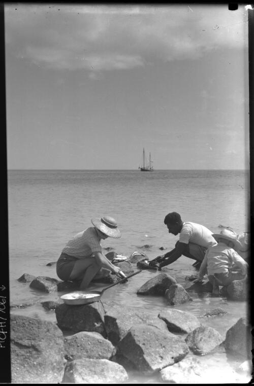 [Man, woman and child, with fresh fish and frypan, boat in distance, 1] [picture] : [Manly, New South Wales] / [Frank Hurley]