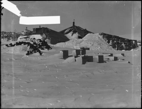 The AAE Main Base Hut at Cape Denison, Adelie Land, with a man on the roof, a mound of snow and boxes, Australasian Antarctic Expedition, approximately 1912 [picture] / Frank Hurley