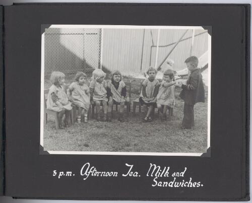 [Photograph of children sitting and standing outside, at afternoon tea, with  milk and sandwiches] [picture]