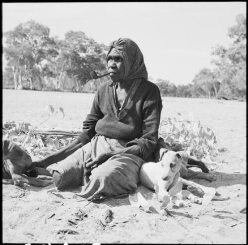 Aboriginal Australian woman smoking a pipe and sitting with two dogs, Alice Springs region, Northern Territory, 1961 / Michael Terry