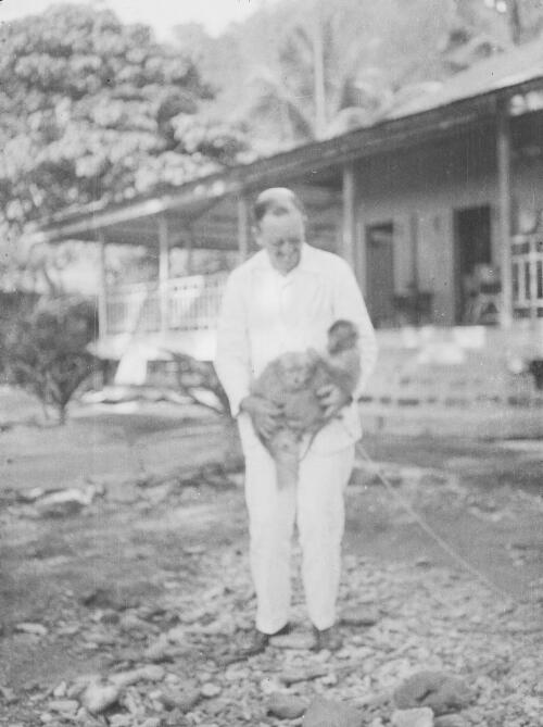 A man holding several monkeys standing in the garden in front of a house, Christmas Island, approximately 1927