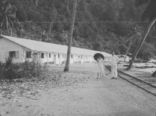 A woman, a man and a small dog standing in front of wooden buildings and railway tracks with dougout canoes lying on the seashore in the background, Christmas Island, approximately 1927