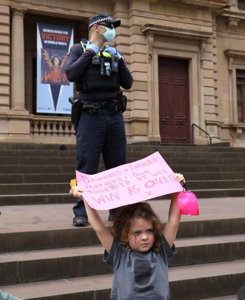 A young girl holding a placard on the steps of the Old Treasury Building, with a police officer standing behind her, during Women's March4Justice rally, Treasury Gardens, Melbourne, 2021 / Leigh Henningham