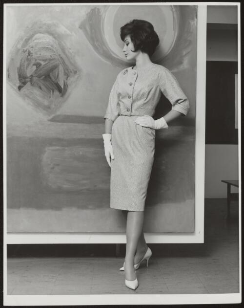 Model wearing a buttoned three quarter sleeve top and knee length pencil skirt standing in front of a painting / Athol Shmith