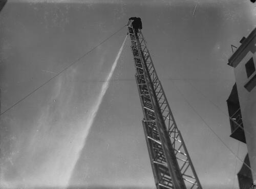 Fireman operating a fire hose at the top of a ladder extended from a fire engine, Newcastle region?, New South Wales, approximately 1950, 2