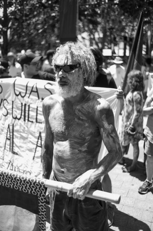 An Aboriginal Australian man with body paint holding a banner, and protestors in the background, during the day of mourning march on Australia Day, Canberra, 26 January 2018 / Sean Davey