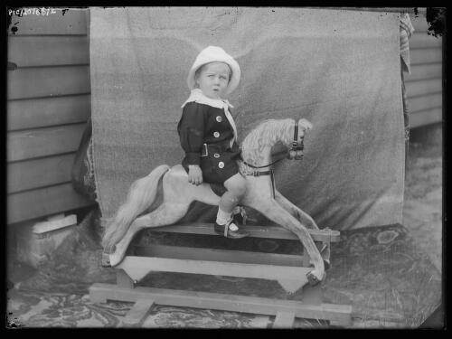 Young boy wearing a coat and a hat sitting on a rocking horse, 1