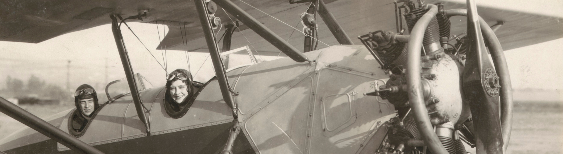 A black and white image of two people sitting in a 1920s style aeroplane while grounded.