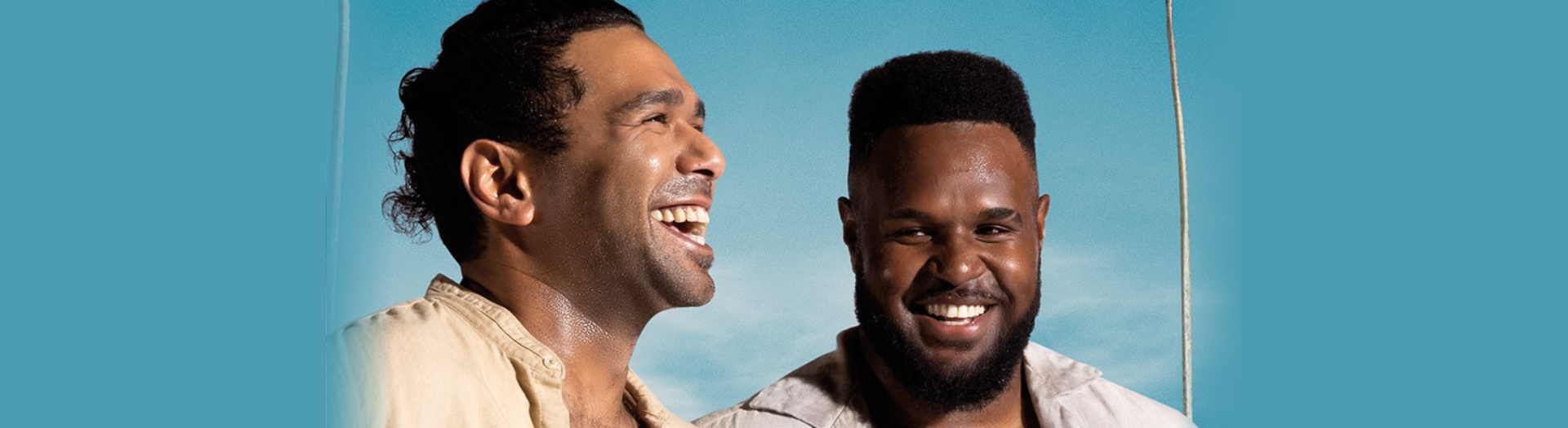 Two men smiling and laughing in front of a blue sky and palm tree.