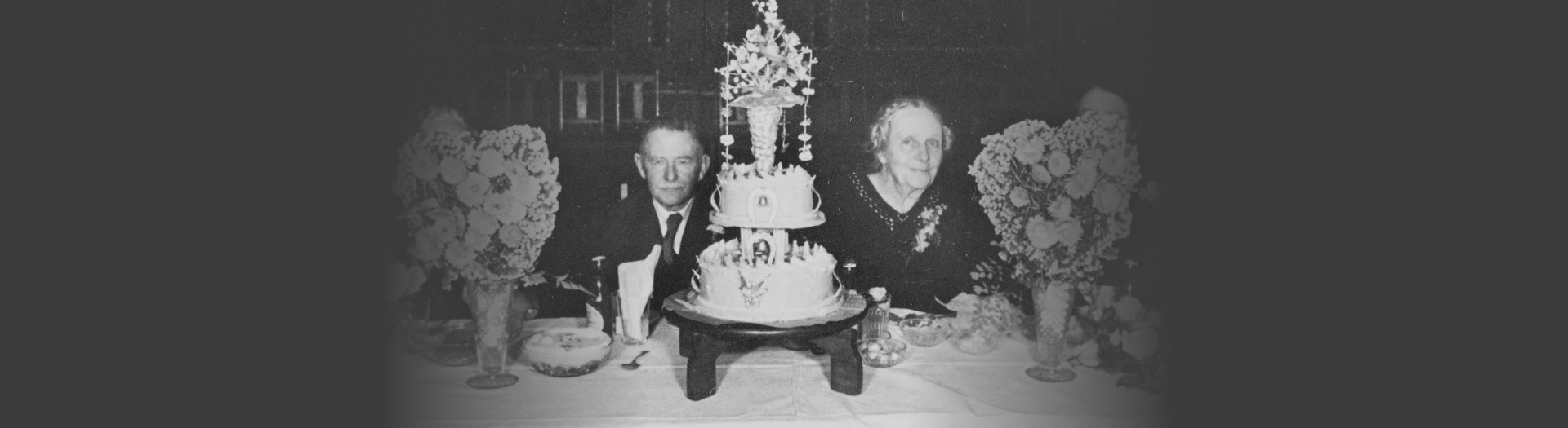 Husband and wife sitting at a table with a large cake and vases of flowers in front of them