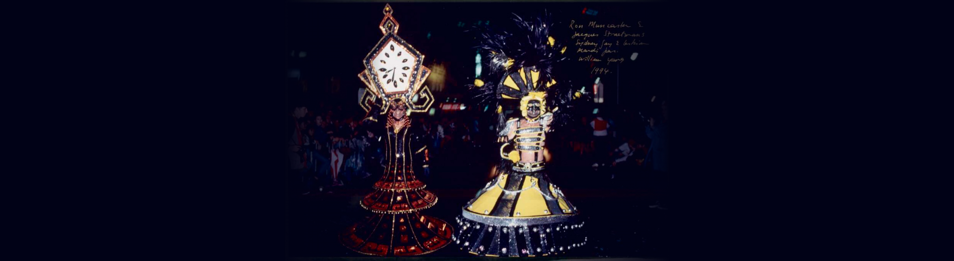 Two men in elaborate costumes, one as an antique clock and the other with yellow and black stripes, walking down a street during a parade 