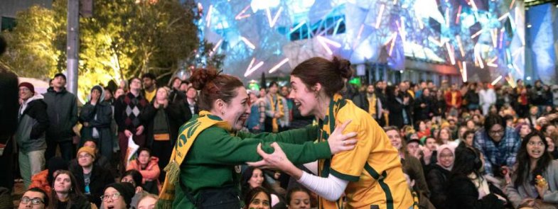 Two woman in green and gold clothing embracing excitedly in front of a crowd.