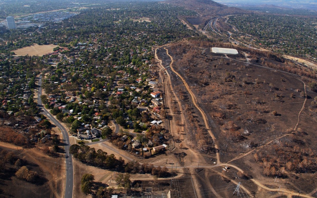 An aerial shot of a city suburb. A hill surrounding the suburb is scarred black from a large fire.